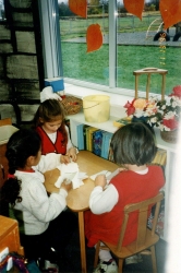 Three girls sitting at table by window working with paper. Orange leaf decals on window. C. Nov 1996