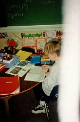 Young boy at table, writing. Large white blur on right side of photograph.