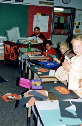 Kids at desks working on halloween decorations, teacher in vampire costume at back.