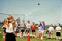 Kids in baseball diamond, orange cones on ground, two girls throwing water balloon(?) to each other. Have faces painted.