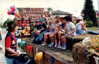 Meadowland Country Fair float in parade. Children sitting on hay barrels atop float, being addressed by woman on left on sidewalk. C.1990